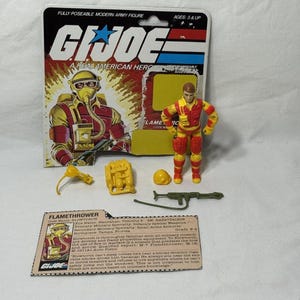 May include: A G.I. Joe Flame Thrower action figure with accessories, including a helmet, backpack, and weapon, is displayed in front of its original packaging. The packaging features the G.I. Joe logo and a character illustration.