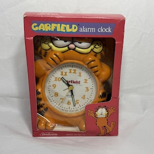 Puede incluir: Reloj despertador Garfield naranja en su caja roja original. La esfera del reloj es blanca con números y manecillas negras. El reloj tiene la forma de Garfield, el gato de dibujos animados, y la caja tiene el logotipo de Garfield.