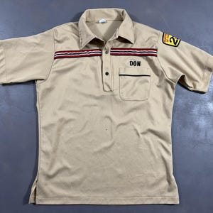 May include: A tan short-sleeved polo shirt with a collar and a pocket. The shirt features a red and black stripe design across the chest and the name "DON" embroidered above the pocket. A patch is on the right sleeve.