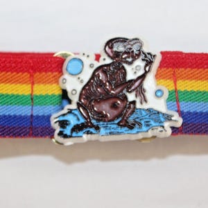 Könnte beinhalten: Ein regenbogenfarbener elastischer Gürtel mit einer Metallschnalle, die eine Comicfigur mit brauner Haut und blauen Augen zeigt. Die Figur hält ein langes, dünnes Objekt in der Hand.