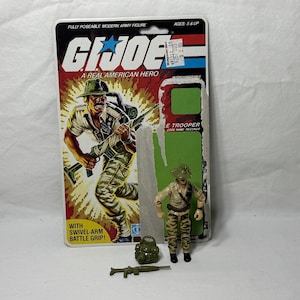 Puede incluir: Una figura de acción vintage de G.I. Joe, aún en su embalaje original. El embalaje presenta una ilustración dinámica de un soldado y el texto "G.I. Joe: A Real American Hero." La figura es un "E Trooper" con una mochila verde y un rifle.