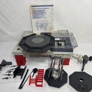 Può includere: Un set da gioco G.I. Joe Tactical Battle Platform con una base grigia e rossa, una piattaforma ottagonale centrale, un centro di controllo in plastica trasparente, un lanciamissili con tre missili, varie armi in plastica nera, scale e un manuale.