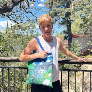 Può includere: Una borsa tote blu con un'illustrazione di procione, montagne e alberi. La borsa ha spallacci blu ed è tenuta da una persona con una canotta bianca. Lo sfondo comprende alberi e un ponte di legno.