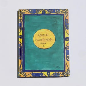 Puede incluir: Un libro de tapa dura titulado "ANIMAL PAINTINGS Volume 1" con una cubierta verde azulado y un círculo amarillo en el centro. El libro tiene un borde decorativo dorado y azul. El nombre del autor, Mae Martin, está impreso en la parte inferior.