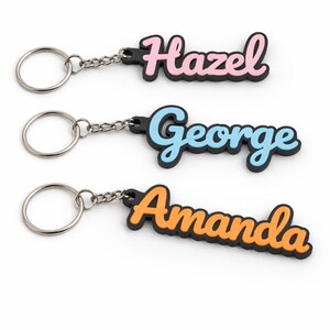 May include: Three personalized keychains with names: "Hazel" in pink, "George" in blue, and "Amanda" in orange. Each name is in a cursive font with a black outline, attached to a silver keyring.