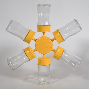 May include: A unique spice rack with six clear glass jars, each with a yellow lid, arranged around a central yellow hub. The jars are evenly spaced, creating a star-like design. The rack is designed for storing spices.