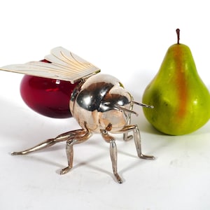 Puede incluir: Naturaleza muerta caprichosa con una escultura de insecto plateado, una esfera de cristal roja y una pera verde. El insecto tiene patas, antenas y alas detalladas. La pera tiene una franja roja vertical.