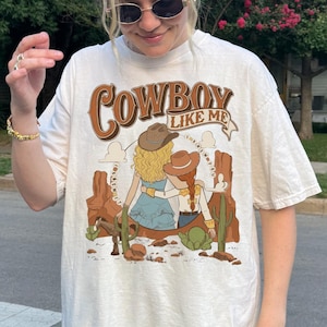 May include: An off-white short-sleeve t-shirt featuring a graphic print with the text "COWBOY LIKE ME" above an illustration of two figures in cowboy hats sitting in a desert landscape with cacti, a horse, and rock formations in shades of brown.