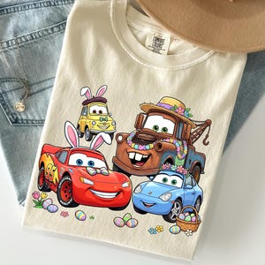 Puede incluir: Camiseta color crema con dibujos de coches de dibujos animados decorados para Semana Santa. El dise&ntilde;o incluye un coche de carreras rojo, un coche deportivo azul, una gr&uacute;a marr&oacute;n y un coche amarillo, todos con orejas de conejito de Pascua y decoraciones florales. Huevos de Pascua y flores.