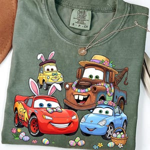 May include: Green t-shirt featuring cartoon cars with bunny ears and Easter eggs. The design includes a red car with the number 95, a yellow car, a brown tow truck, and a blue car. The shirt has a Comfort Colors tag.