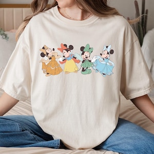 Puede incluir: Camiseta color crema con un gr&aacute;fico de cuatro personajes de dibujos animados con vestidos coloridos. Los personajes est&aacute;n dispuestos en fila, cada uno con un atuendo y una pose &uacute;nicos. La camiseta es de corte holgado.