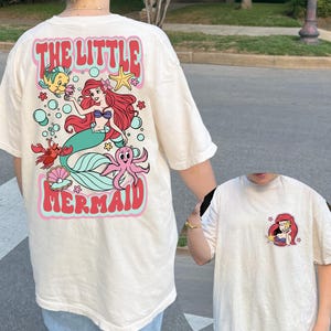 Pu&ograve; includere: T-shirt color crema con grafica di Ariel de La Sirenetta. Il retro della maglietta mostra Ariel, Flounder, Sebastian e un polpo, con le parole "THE LITTLE MERMAID" in un font retr&ograve;. Una grafica pi&ugrave; piccola di Ariel &egrave; sul davanti.