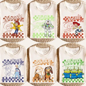 May include: Six cream-colored t-shirts featuring Toy Story characters. Each shirt has a different character design: Woody, Buzz, Jessie, Bo Peep, Slinky, and Aliens. Each design includes the character's name and the year 1995.