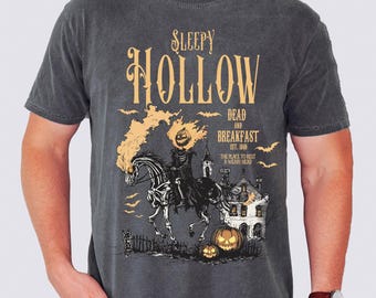 May include: Dark gray t-shirt featuring a Halloween-themed graphic. The design includes the text "Sleepy Hollow Dead and Breakfast" with an illustration of a headless horseman riding a horse, a haunted house, and pumpkins.