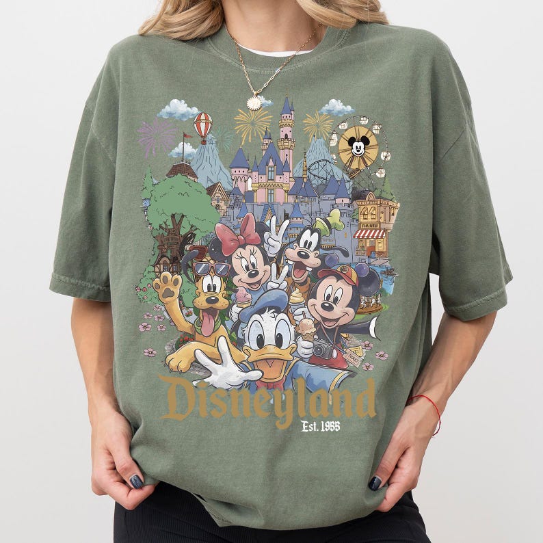 May include: An olive green t-shirt featuring a colorful graphic print of Disney characters including Mickey Mouse, Minnie Mouse, Donald Duck, Goofy, and Pluto in front of a castle. The text Disneyland Est. 1955 is printed in gold at the bottom.