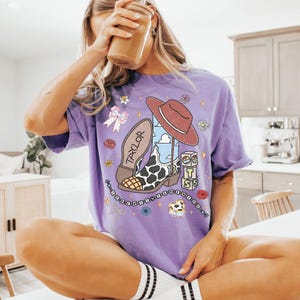 May include: A purple short-sleeve t-shirt featuring a graphic print of a cowboy boot with a cow print pattern, a brown hat, the name TAYLOR, the number 13, and blocks spelling BTS. The design includes small flowers, a bow, and a beaded string.