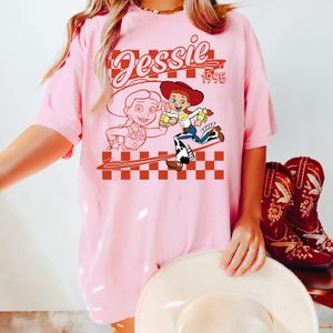 May include: Pink t-shirt featuring a graphic of the character Jessie from the movie Toy Story, with the text "Jessie 1995". The design includes a checkered pattern and a cartoon illustration of Jessie. A pair of red cowboy boots are visible.