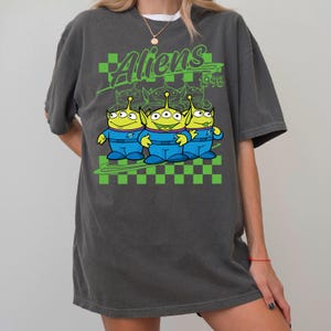 May include: Dark gray t-shirt featuring a graphic of three green aliens in blue uniforms with the text "Aliens 1995" in green. The design includes a green and black checkered pattern.