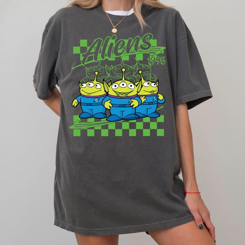 May include: Dark gray t-shirt featuring a graphic of three green aliens in blue uniforms with the text "Aliens 1995" in green. The design includes a green and black checkered pattern.