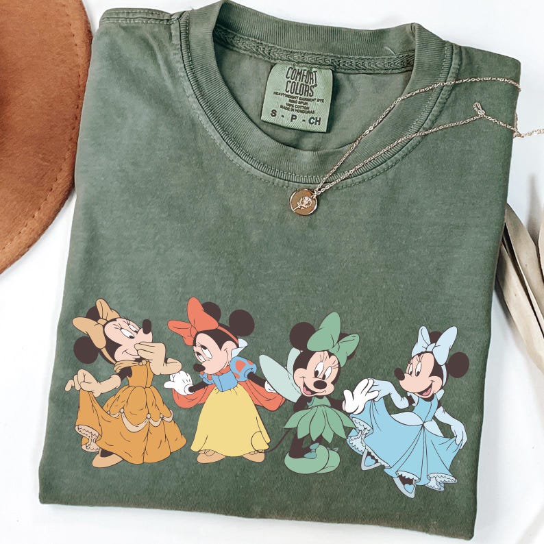 Puede incluir: Camiseta verde oliva Comfort Colors con cuatro personajes de Minnie Mouse con vestidos de princesa. La camiseta tiene cuello redondo y mangas cortas. Un collar dorado con un colgante est&aacute; sobre la camiseta.