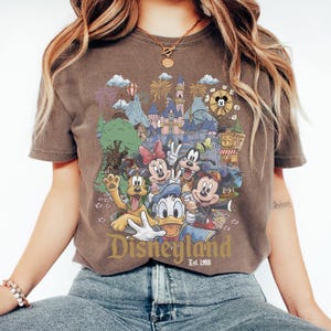 May include: A brown short-sleeve t-shirt featuring a graphic print of Disney characters including Mickey Mouse, Minnie Mouse, Donald Duck, Goofy, and Pluto in front of a castle. The text Disneyland Est. 1955 is printed in gold at the bottom.