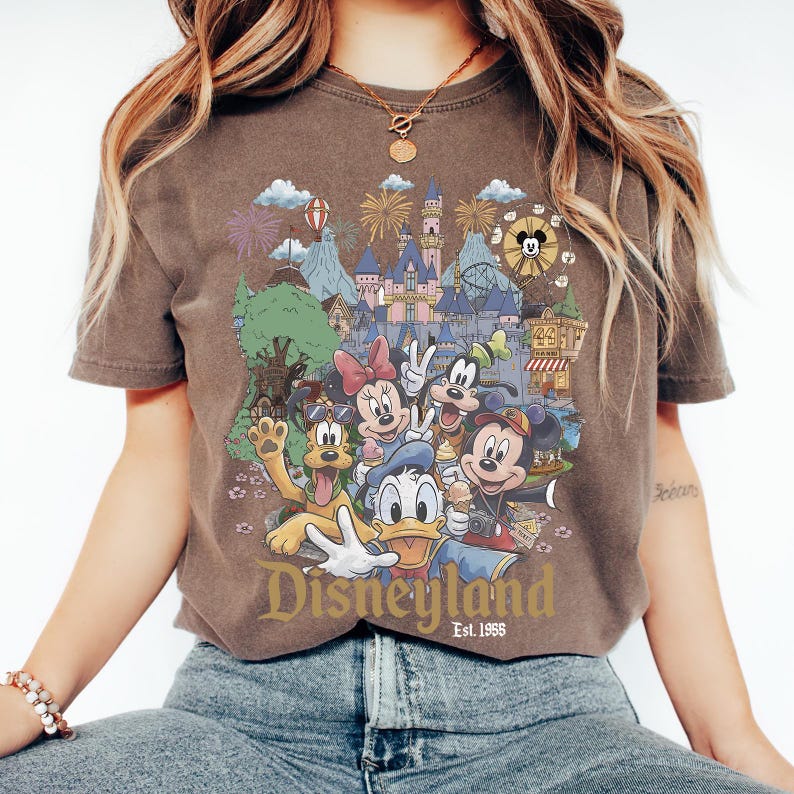 May include: A brown short-sleeve t-shirt featuring a graphic print of Disney characters including Mickey Mouse, Minnie Mouse, Donald Duck, Goofy, and Pluto in front of a castle. The text Disneyland Est. 1955 is printed in gold at the bottom.