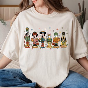 May include: Cream-colored t-shirt featuring a festive design with cartoon characters dressed as nutcrackers. The characters include Mickey Mouse, Minnie Mouse, Donald Duck, and Goofy, with colorful details and Christmas lights.