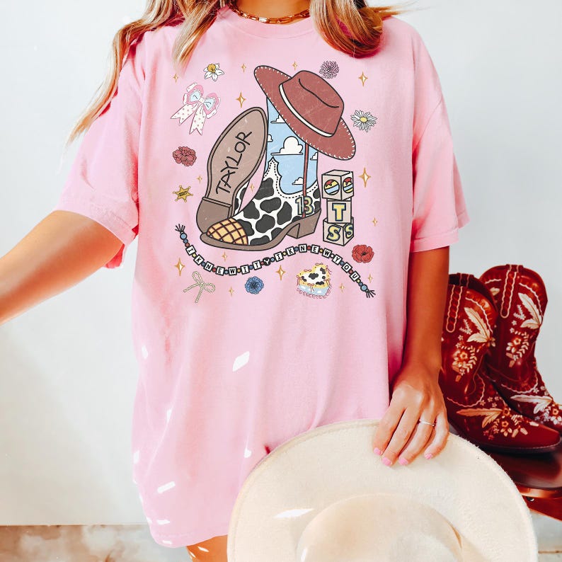 May include: A light pink t-shirt featuring a graphic print of a brown cowboy hat, a cow-print cowboy boot with the number 13, a brown boot sole labeled TAYLOR, three stacked blocks with the letters T, S, and a heart, and a beaded string with text.