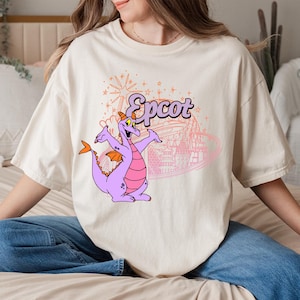 May include: Cream-colored t-shirt featuring a purple dragon character with orange wings and a pink belly. The word "Epcot" is above the dragon in a stylized font, with a pink outline of a theme park in the background.