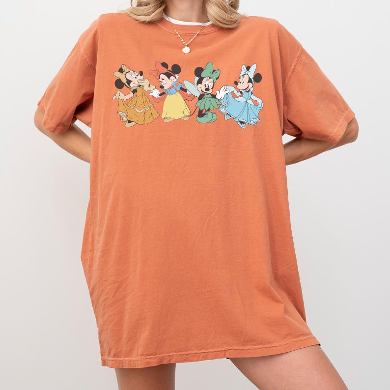 Puede incluir: Una camiseta naranja con un gr&aacute;fico de cuatro personajes de Minnie Mouse con diferentes vestidos. La camiseta tiene mangas cortas y un corte holgado. El gr&aacute;fico es de colores brillantes.