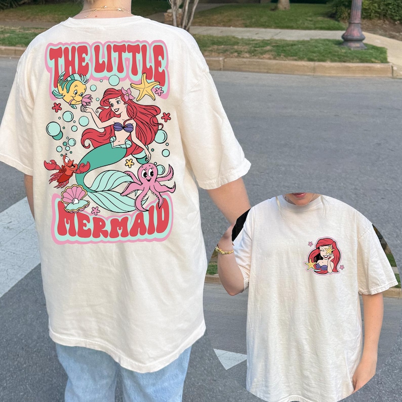 Pu&ograve; includere: T-shirt color crema con grafica di Ariel de La Sirenetta. Il retro della maglietta mostra Ariel, Flounder, Sebastian e un polpo, con le parole "THE LITTLE MERMAID" in un font retr&ograve;. Una grafica pi&ugrave; piccola di Ariel &egrave; sul davanti.