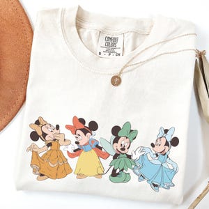 Puede incluir: Camiseta color crema con cuatro personajes de Minnie Mouse con vestidos de princesa. La camiseta tiene cuello redondo y mangas cortas. Un collar dorado con un colgante descansa sobre la camiseta. La etiqueta dice "Comfort Colors".