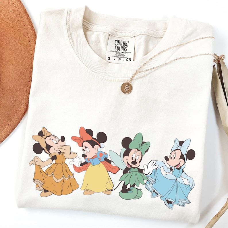 Puede incluir: Camiseta color crema con cuatro personajes de Minnie Mouse con vestidos de princesa. La camiseta tiene cuello redondo y mangas cortas. Un collar dorado con un colgante descansa sobre la camiseta. La etiqueta dice "Comfort Colors".