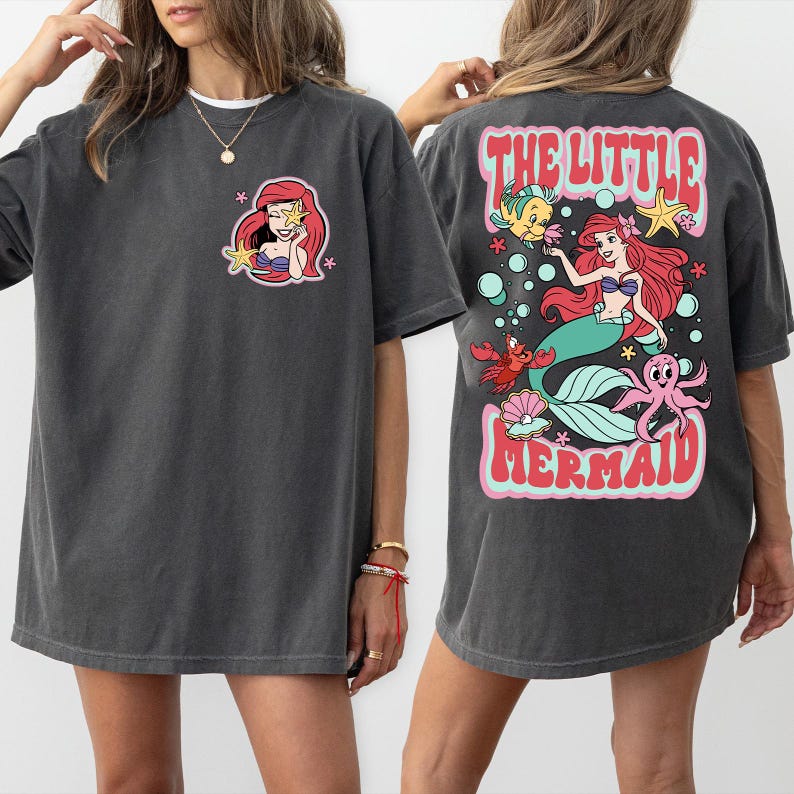Pu&ograve; includere: T-shirt grigio scuro con una grafica a cartoni animati di una sirena sul davanti. Il retro della maglietta mostra la scritta "THE LITTLE MERMAID" con una sirena e creature marine, tra cui una stella marina.