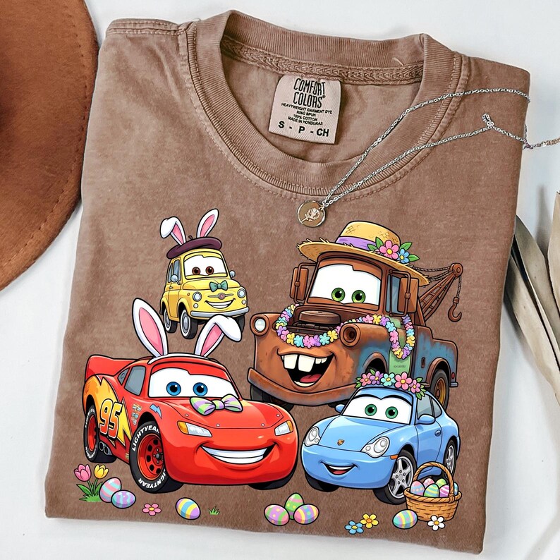 Puede incluir: Camiseta marr&oacute;n con dibujos de coches con orejas de conejo y huevos de Pascua. La camiseta tiene el texto "Comfort Colors" en la etiqueta. Un collar de plata est&aacute; drapeado sobre la camiseta.