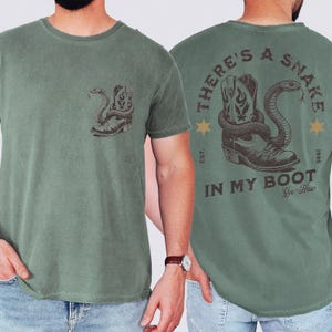May include: A sage green short-sleeve t-shirt featuring a small graphic on the front left chest and a large graphic on the back. Both graphics depict a cowboy boot with a snake coiled around it. The back text reads, "THERE'S A SNAKE IN MY BOOT".