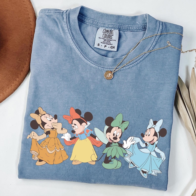 Puede incluir: Una camiseta azul Comfort Colors con cuatro personajes de Minnie Mouse con vestidos coloridos. Un collar dorado con un colgante descansa sobre la camiseta. La camiseta est&aacute; doblada sobre una superficie blanca.