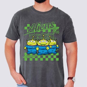 May include: Dark gray t-shirt featuring a graphic of three green aliens with three eyes each, wearing blue uniforms. The word "Aliens" is printed above the aliens in green, along with the year "1995". The design includes a green and black checkered pattern.
