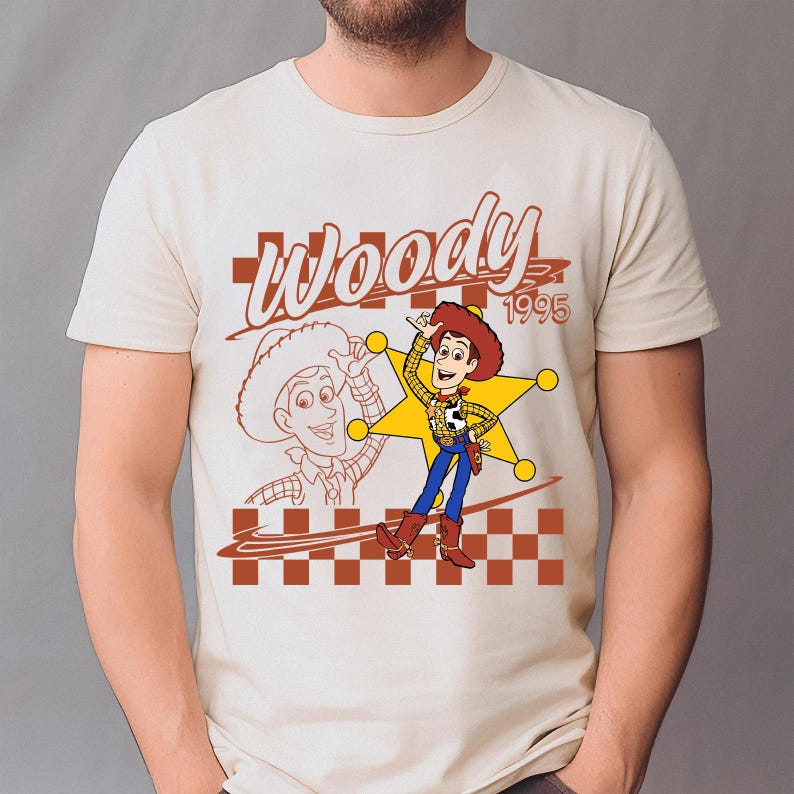 May include: Cream-colored t-shirt featuring a graphic of Woody from Toy Story. The design includes a cartoon Woody character, a sheriff's star, and the word "Woody" with the year "1995". A brown and white checkered pattern is at the bottom.