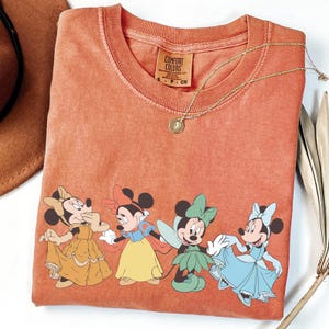 Puede incluir: Camiseta naranja Comfort Colors con cuatro ratones de dibujos animados con vestidos coloridos. Un collar dorado con un colgante descansa sobre la camiseta. Un sombrero marr&oacute;n y hojas secas est&aacute;n en el fondo.