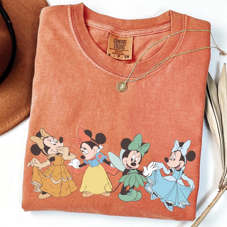 Puede incluir: Camiseta naranja Comfort Colors con cuatro ratones de dibujos animados con vestidos coloridos. Un collar dorado con un colgante descansa sobre la camiseta. Un sombrero marr&oacute;n y hojas secas est&aacute;n en el fondo.