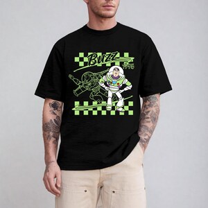 May include: Black t-shirt featuring a neon green graphic of Buzz Lightyear from Toy Story. The design includes the word "Buzz" and the year "1995" on a checkered background. The shirt is a crew neck style.