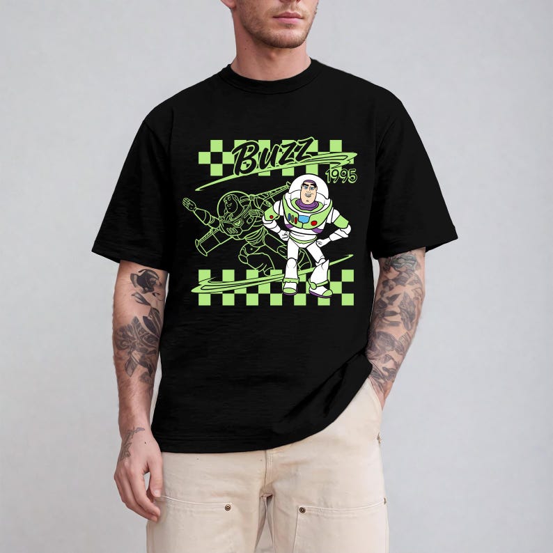 May include: Black t-shirt featuring a neon green graphic of Buzz Lightyear from Toy Story. The design includes the word "Buzz" and the year "1995" on a checkered background. The shirt is a crew neck style.