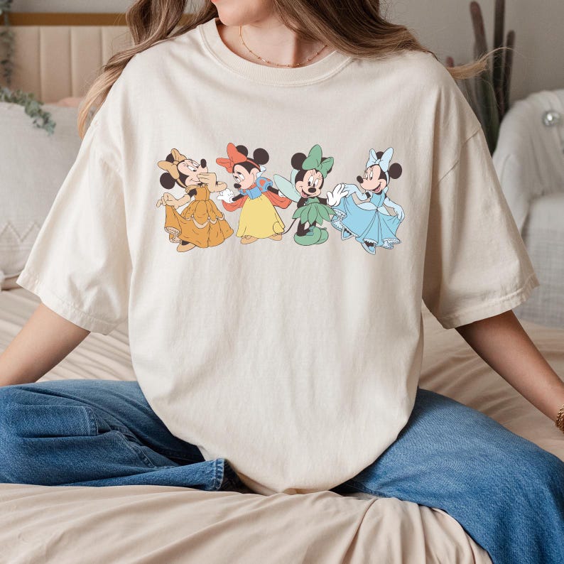 Puede incluir: Camiseta color crema con un gr&aacute;fico de cuatro personajes de dibujos animados con vestidos coloridos. Los personajes est&aacute;n dispuestos en fila, cada uno con un atuendo y una pose &uacute;nicos. La camiseta es de corte holgado.