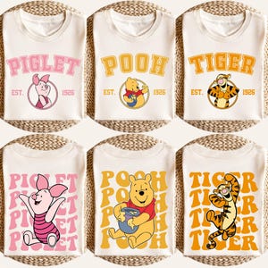 May include: Cream-colored t-shirts featuring cartoon characters Piglet, Pooh, and Tigger. Each shirt has the character's name in a retro font, with "EST. 1926" below. The bottom row repeats the characters' names in a stacked design.