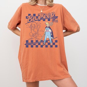 May include: Orange t-shirt featuring a graphic of Bo Peep from Toy Story. The design includes the text "Bo Peep 1995" in blue, along with two illustrations of the character. The shirt has a relaxed fit.