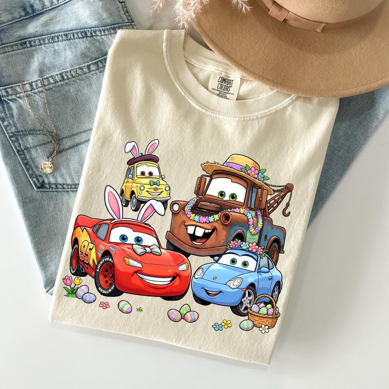 Puede incluir: Camiseta color crema con dibujos de coches de dibujos animados decorados para Semana Santa. El dise&ntilde;o incluye un coche de carreras rojo, un coche deportivo azul, una gr&uacute;a marr&oacute;n y un coche amarillo, todos con orejas de conejito de Pascua y decoraciones florales. Huevos de Pascua y flores.
