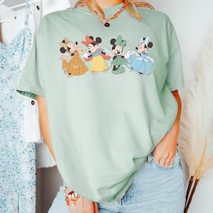 Puede incluir: Camiseta verde claro con un gr&aacute;fico de cuatro personajes de Minnie Mouse con diferentes vestidos. Los personajes de dibujos animados est&aacute;n en fila, cada uno con un vestido de diferente color. La camiseta tiene mangas cortas y cuello redondo.
