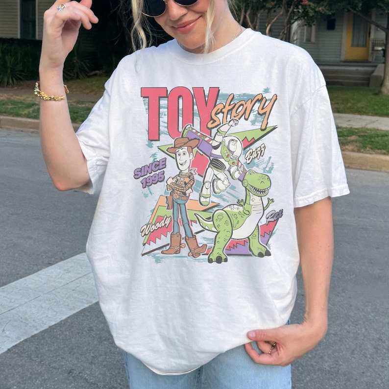 May include: White t-shirt featuring a colorful graphic of characters from the movie "Toy Story." The design includes Woody, Buzz Lightyear, and Rex, with the text "TOY STORY" and "SINCE 1995." The shirt has a vintage, retro style.