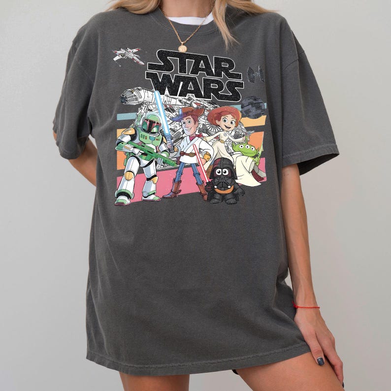 May include: Dark gray Star Wars graphic t-shirt featuring cartoon characters and the Star Wars logo. The design includes a green and white Stormtrooper, Woody, and other characters on a striped background. A casual, oversized fit.